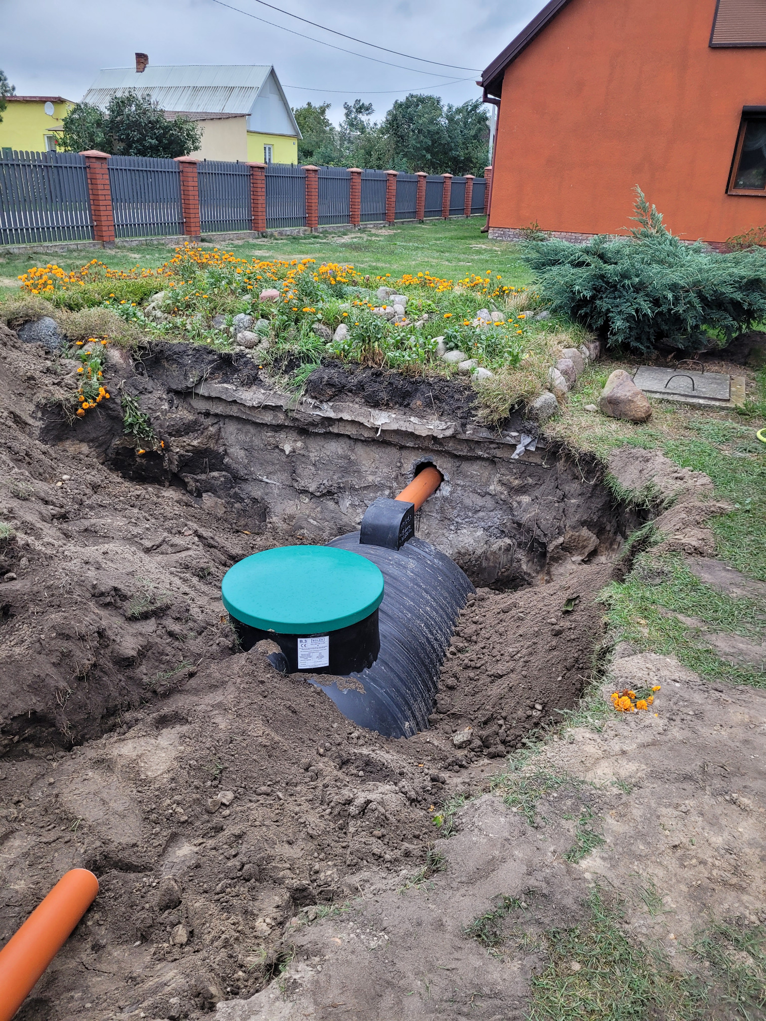 Czarny zbiornik z zieloną pokrywą umieszczony w wykopie ziemnym, widoczny fragment pomarańczowej rury, w tle ogród z kwiatami i zabudowania.