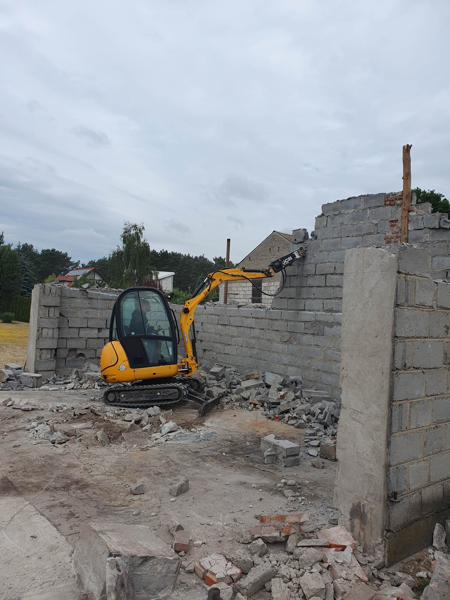 Żółta minikoparka JCB z młotem hydraulicznym w trakcie rozbiórki ściany z szarych bloczków betonowych, w tle widoczne domy i drzewa.