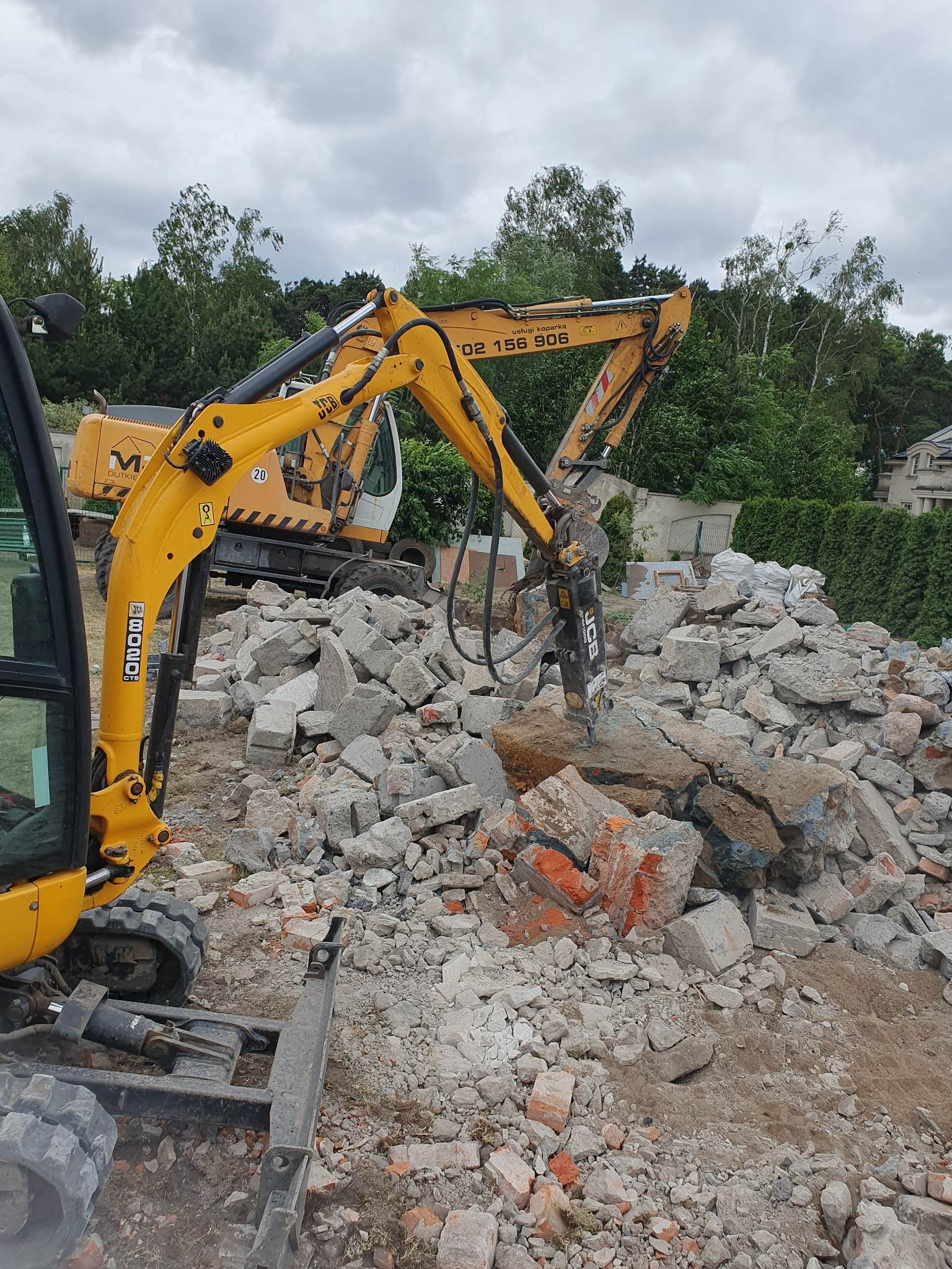 Żółta koparka JCB z młotem hydraulicznym rozbija gruz betonowy i ceglany na placu budowy, w tle widoczna druga koparka i zieleń drzew