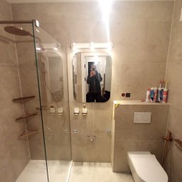 plytkarz. - Wykończona łazienka w beżowych płytkach z kabiną prysznicową typu walk-in z elementami w kolorze miedzianym, wiszącą toaletą i lustrem z podświetleniem. W lustrze odbija się osoba robiąca zdjęcie.