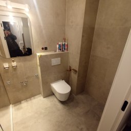 plytkarz. - Wykończona łazienka z beżowymi płytkami, widoczna toaleta podtynkowa, akcesoria łazienkowe w kolorze miedzi, oraz tuby z silikonem na zabudowie stelaża.