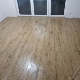 Nowo ułożona, błyszcząca podłoga z paneli imitujących drewno, widoczna przy białych drzwiach balkonowych z żaluzjami.