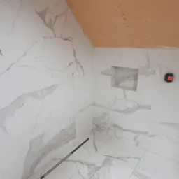 Łazienka w trakcie wykańczania: białe płytki imitujące marmur na ścianach i podłodze, widoczny odpływ liniowy, wnęka w ścianie i fragment sufitu z płyt kartonowo-gipsowych.