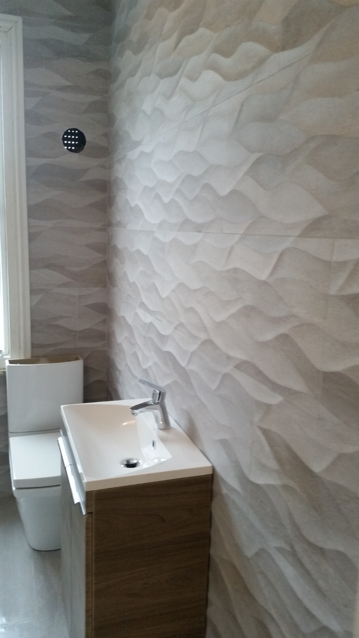 Nowoczesna łazienka z umywalką na szafce z drewnopodobnym wykończeniem i toaletą, ściany wyłożone płytkami z trójwymiarowym wzorem fal, widoczny nawiewnik wentylacyjny.