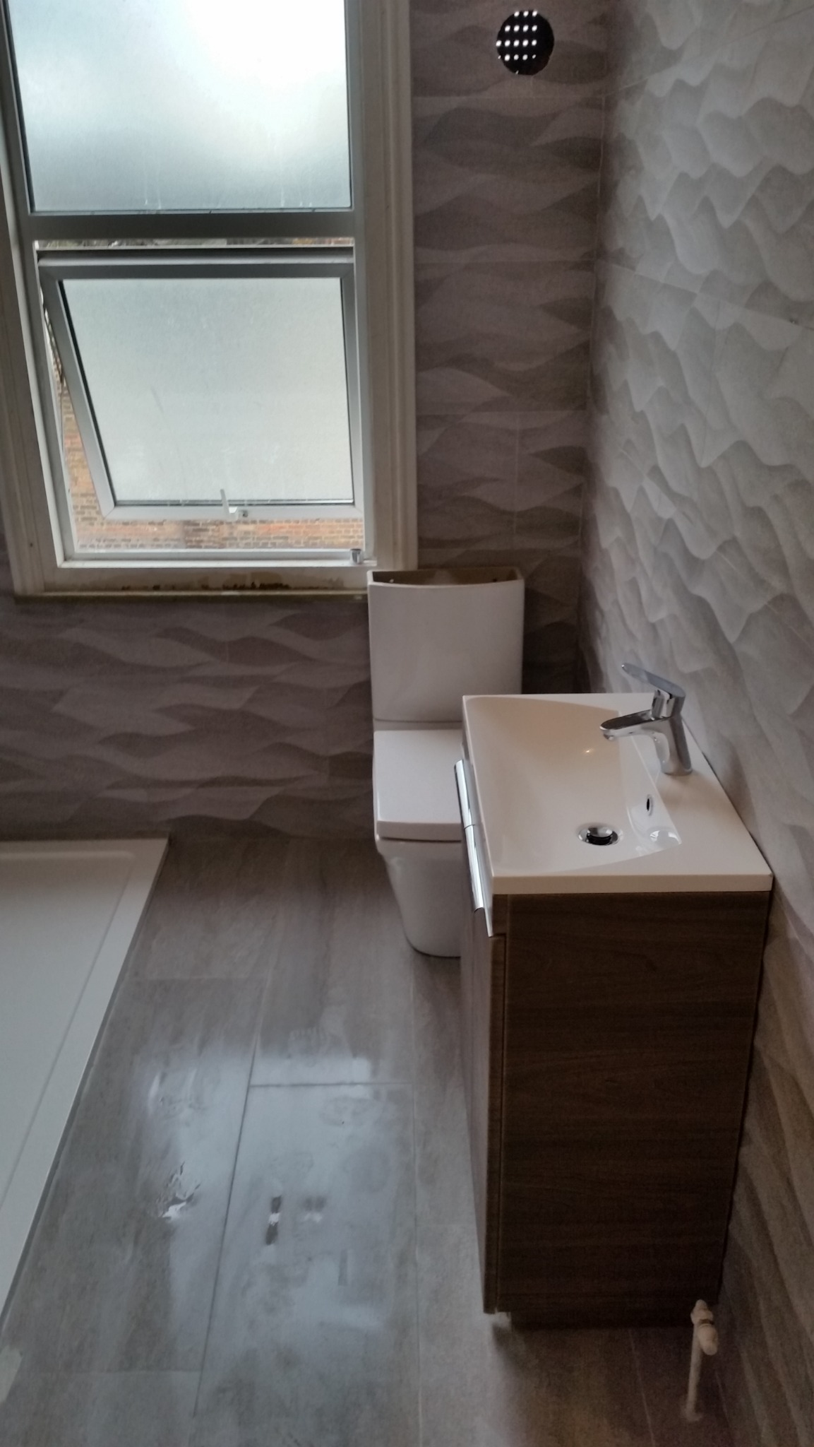 Nowoczesna łazienka po remoncie: umywalka z szafką w kolorze drewna, biała toaleta, szare płytki z falistym wzorem na ścianach i podłodze, okno z uchylną górną częścią.