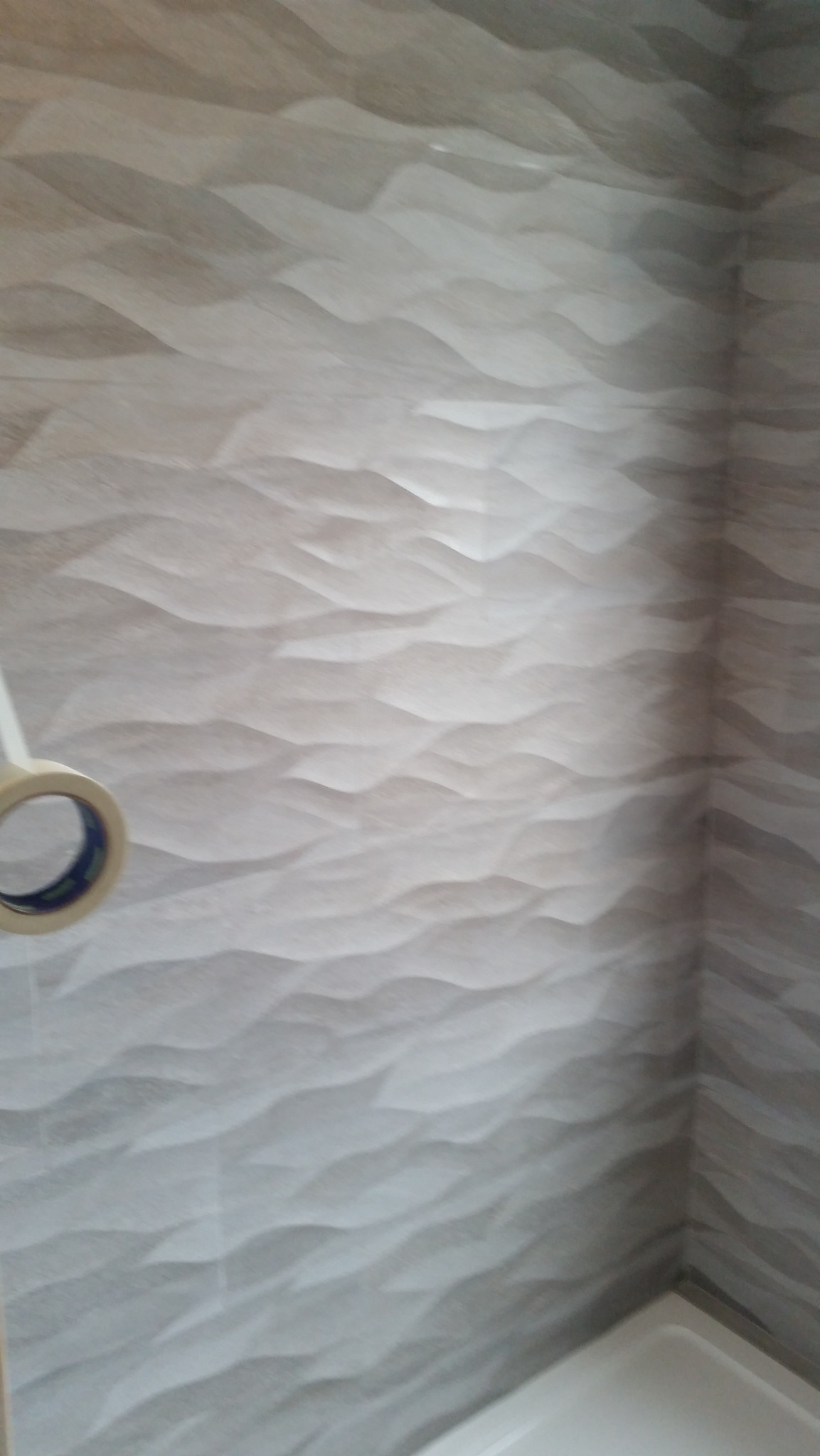 Ściana wyłożona trójwymiarowymi, beżowo-szarymi płytkami ceramicznymi o falistej strukturze w rogu pomieszczenia; widoczny fragment taśmy malarskiej i brodzika.