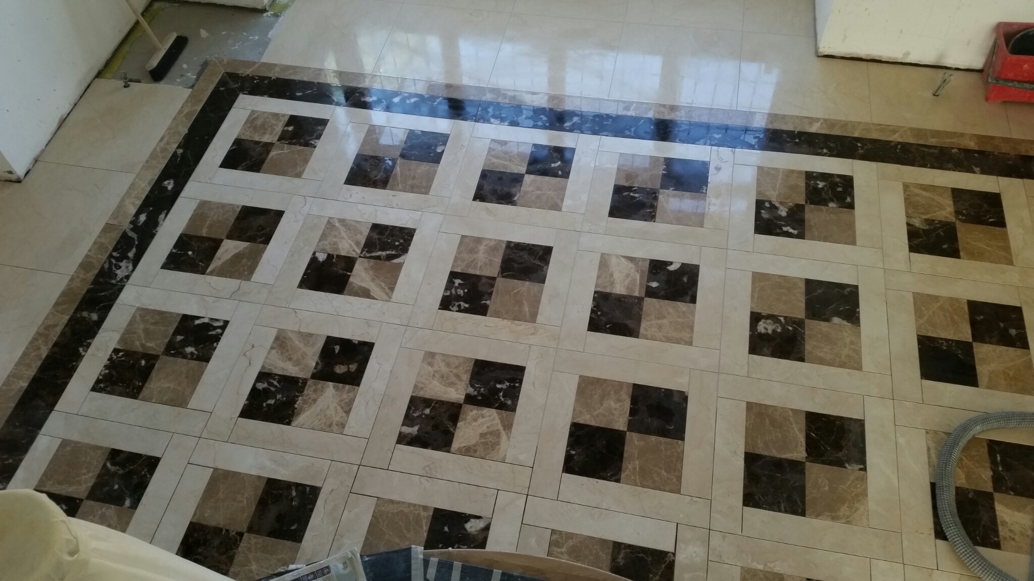 Ułożona podłoga z płytek w kwadraty w odcieniach beżu i brązu z ciemną obwódką, widoczne odbicia światła na powierzchni, fragment odkurzacza w rogu.