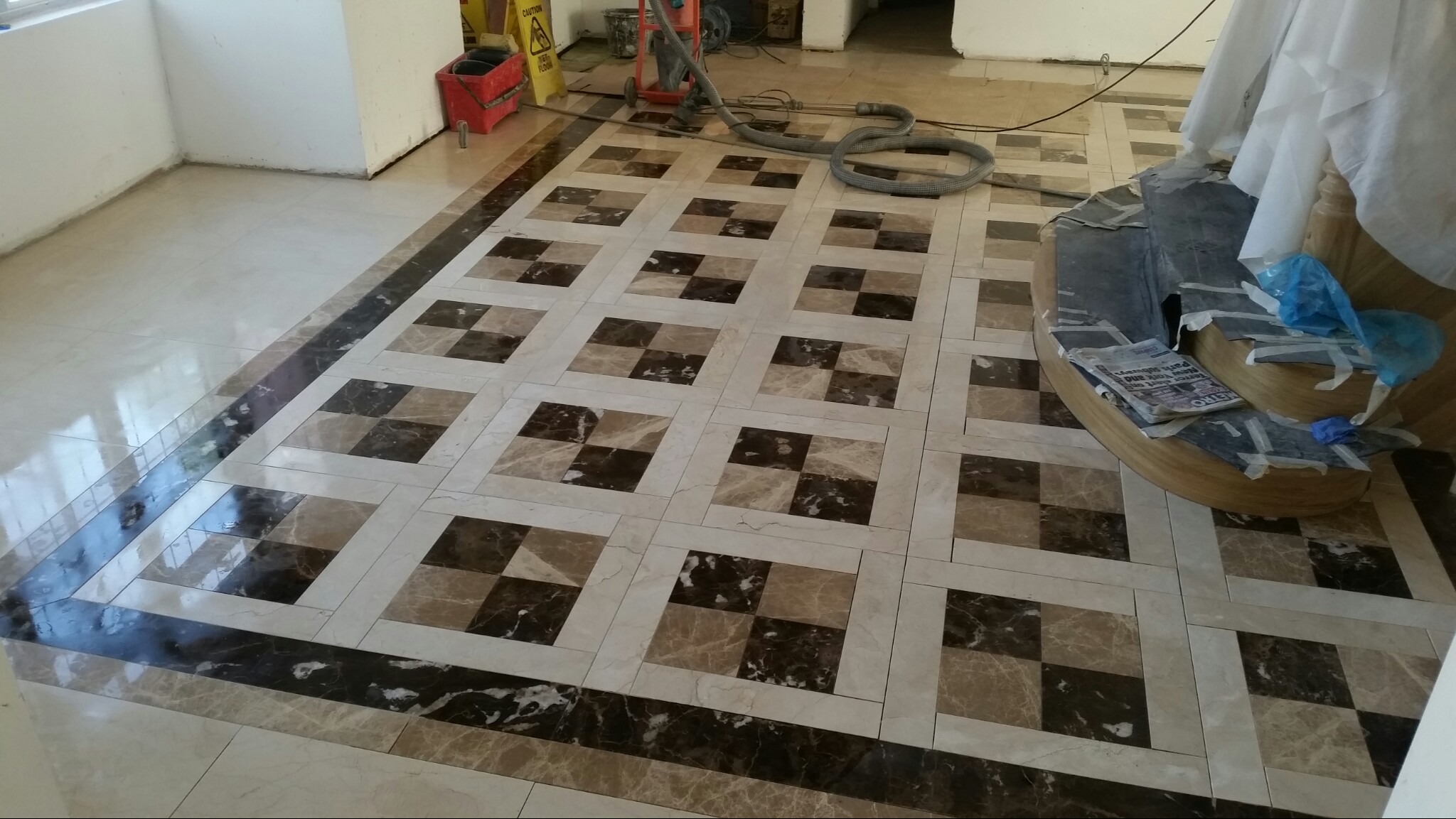 Wzorzysta podłoga z płytek w odcieniach beżu i brązu, ułożona w kwadraty z ciemną bordiurą, widoczne narzędzia i akcesoria do czyszczenia podłóg oraz zabezpieczone schody w tle.