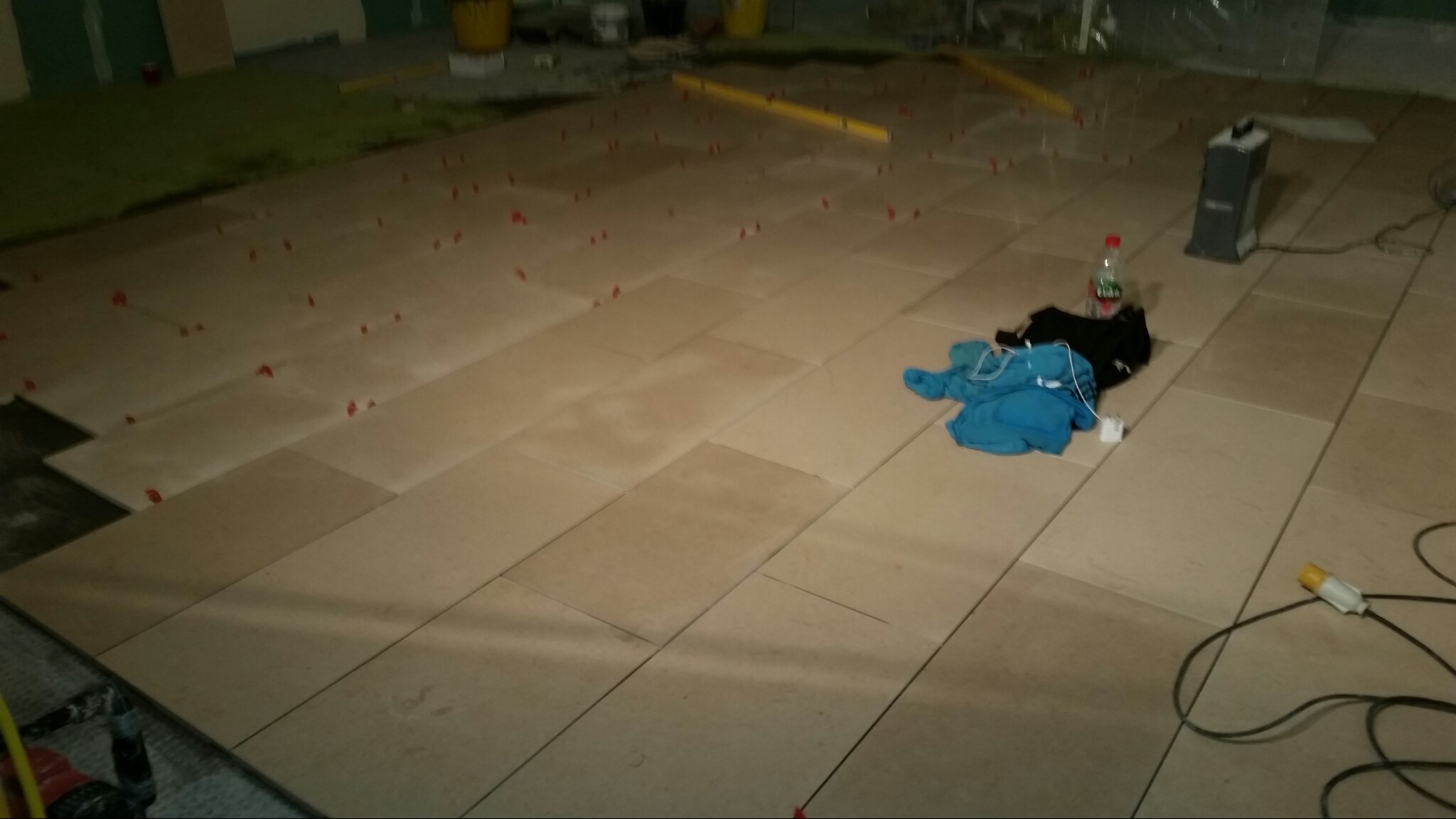 Układanie beżowych płytek podłogowych z użyciem czerwonych krzyżyków dystansowych, widoczne narzędzia, poziomica, odzież robocza i butelka wody.