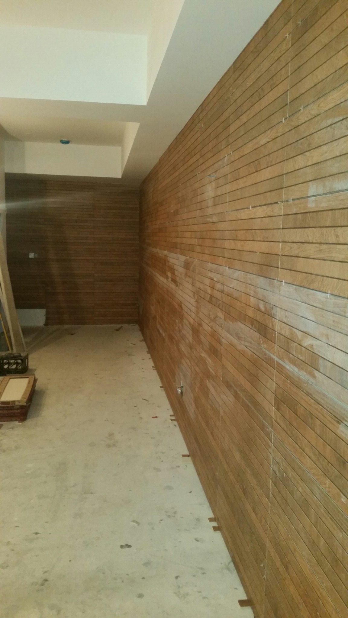 Ściana wykończona drewnopodobnymi płytkami w trakcie montażu, widoczne krzyżyki dystansowe, surowy beton na podłodze i ślady zaprawy.