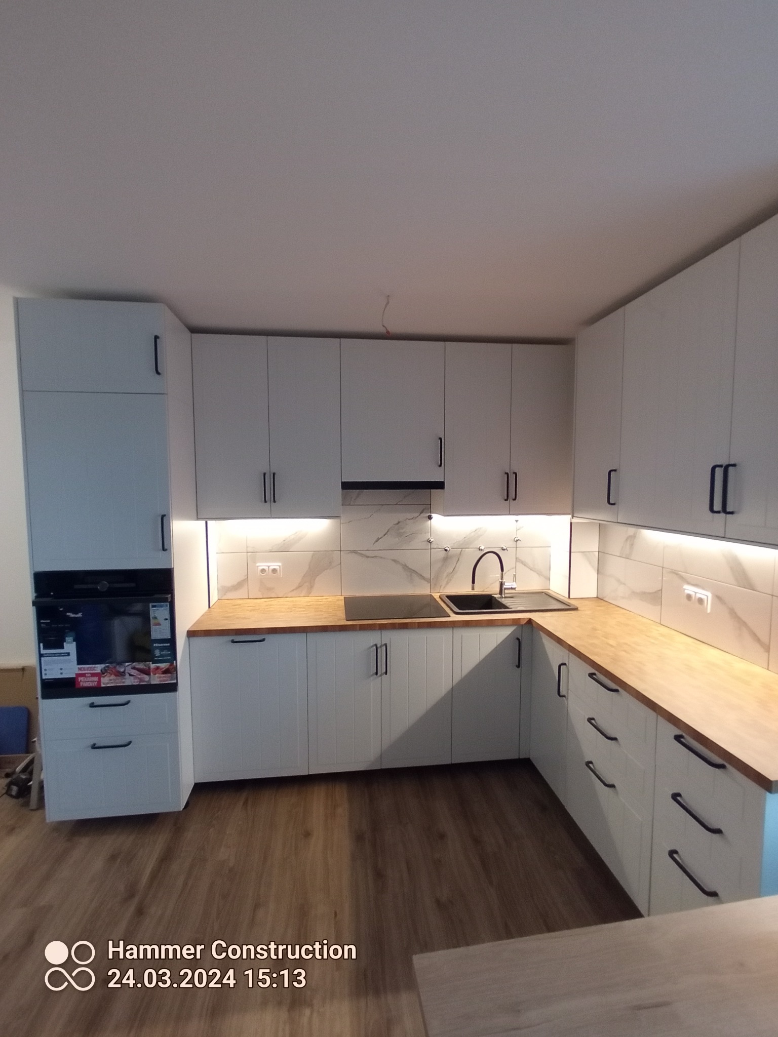 Nowoczesna kuchnia z białymi szafkami z czarnymi uchwytami, drewnianym blatem i marmurową ścianą nad blatem, oświetlona taśmą LED pod szafkami wiszącymi.