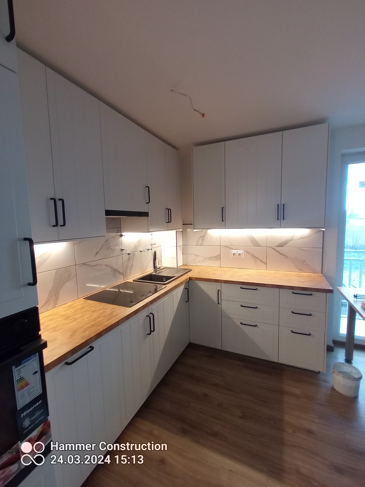 Nowoczesna kuchnia z białymi szafkami z czarnymi uchwytami, drewnianym blatem i marmurowymi płytkami na ścianie, oświetlona taśmami LED pod szafkami wiszącymi.