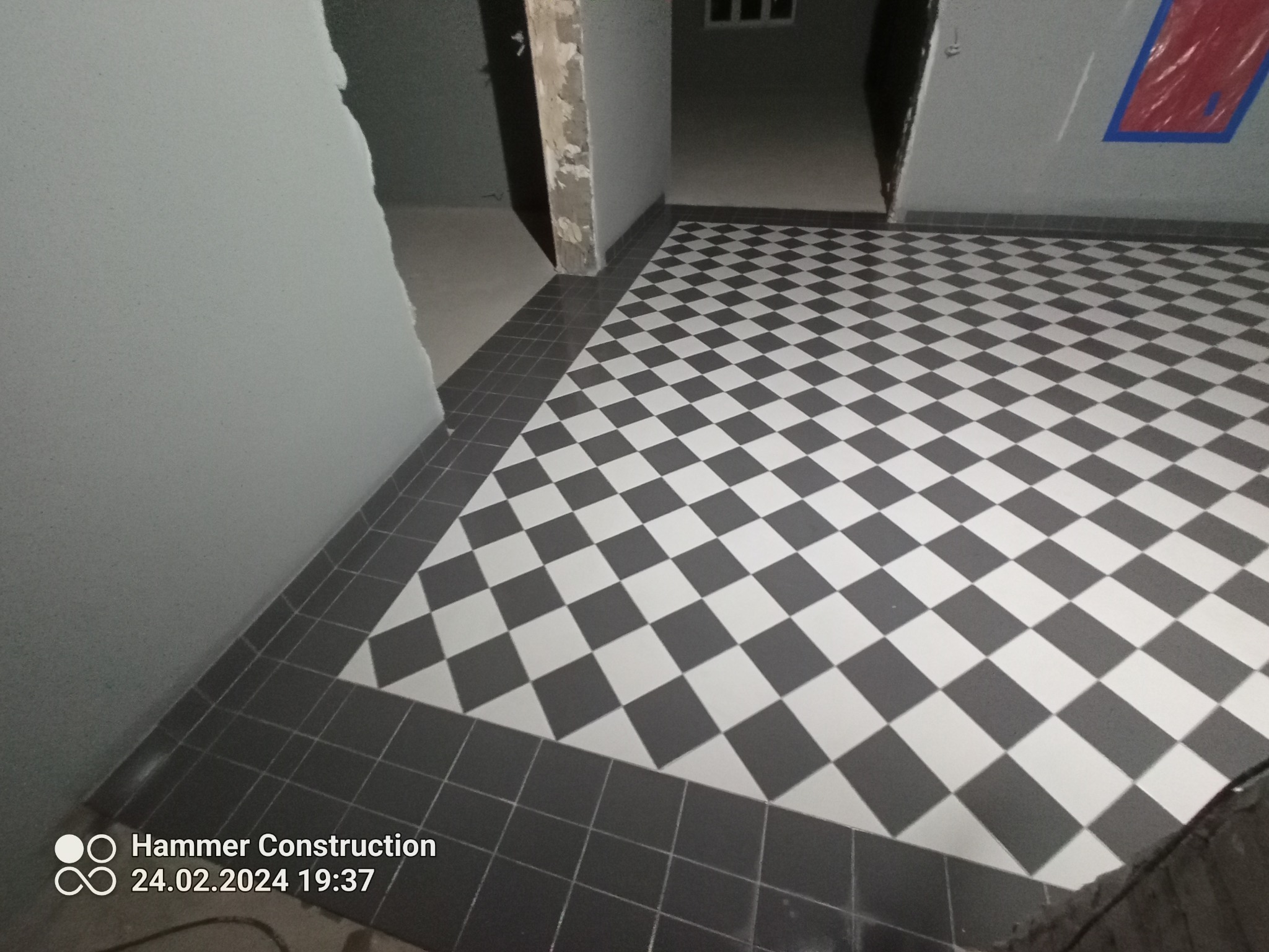 Ułożona podłoga z płytek w szachownicę, czarno-białych, z ciemnoszarą obwódką. Widoczne surowe ściany w trakcie wykończenia i fragment logo firmy w rogu zdjęcia.