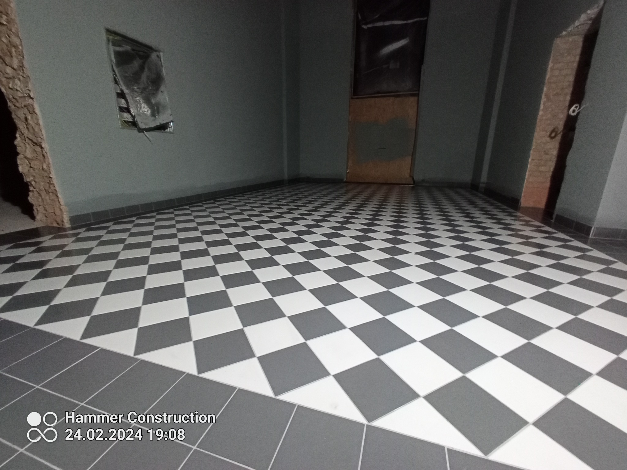 Ukończona podłoga w szachownicę z szarych i białych płytek, widoczne fragmenty ściany w szarym kolorze, z niedokończonym wykończeniem przy futrynie drzwi.