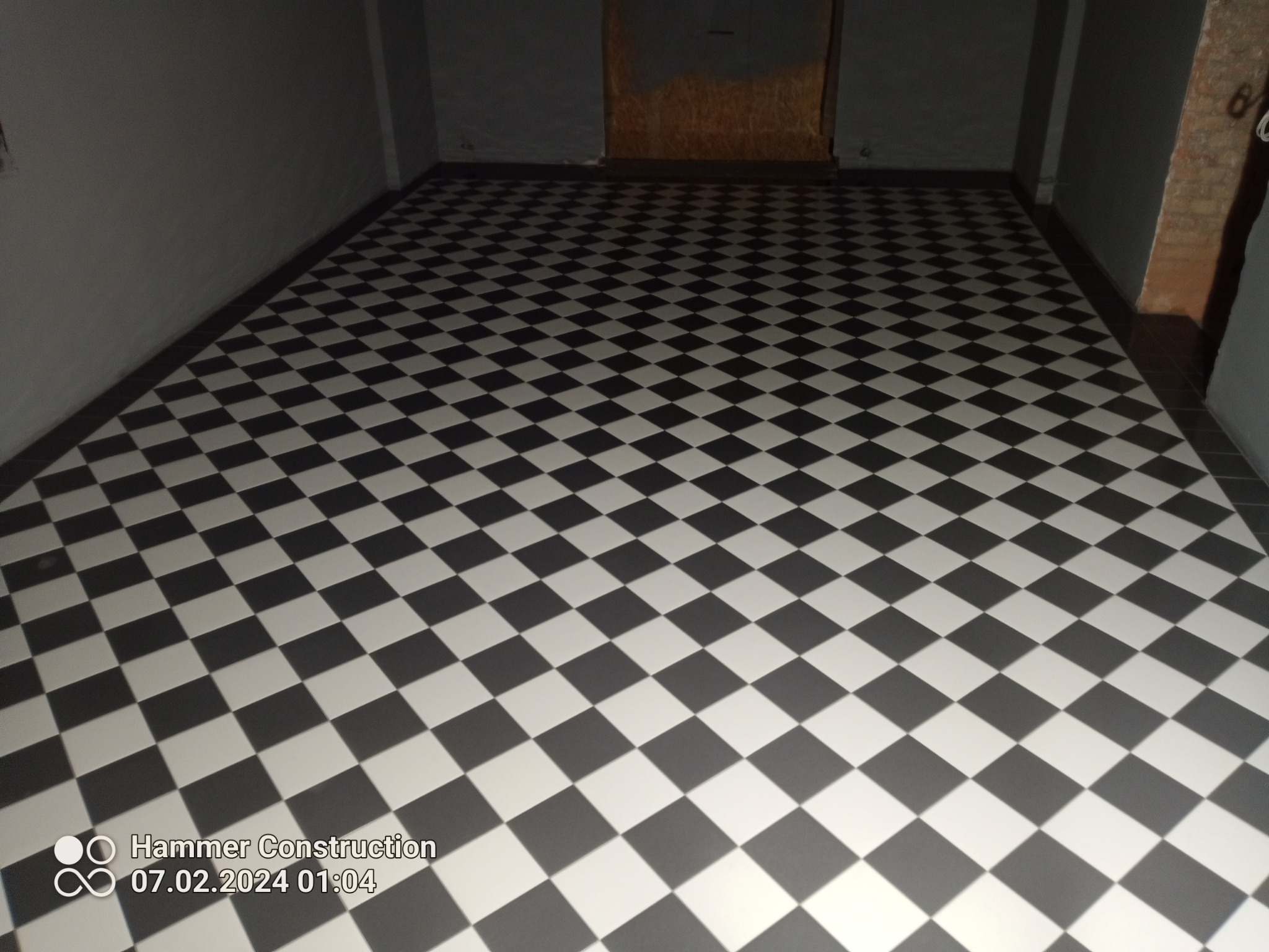 Ułożona podłoga z płytek w szachownicę, czarno-białe płytki ułożone pod kątem w ciemnym pomieszczeniu, widoczne ściany i fragment drzwi.