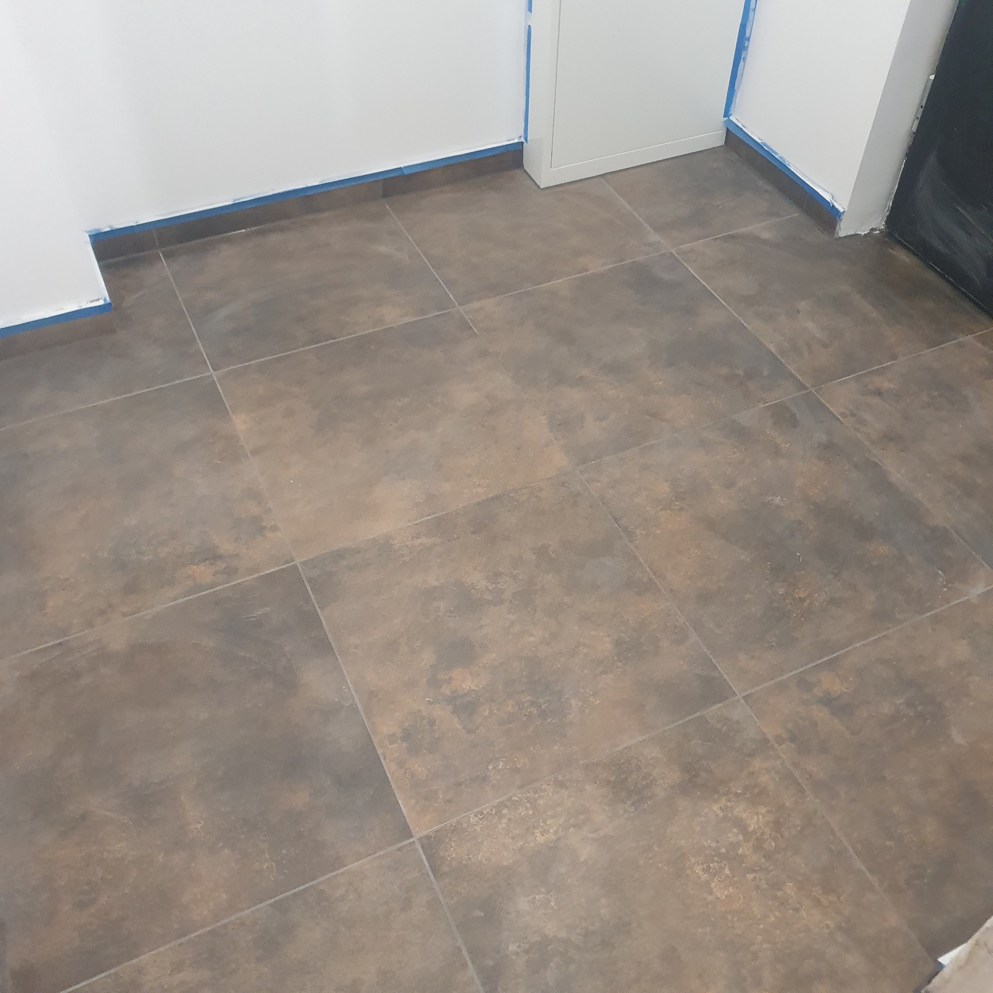 Ułożone, brązowo-szare płytki podłogowe w pomieszczeniu z białymi ścianami, zabezpieczonymi niebieską taśmą malarską przy podłodze.