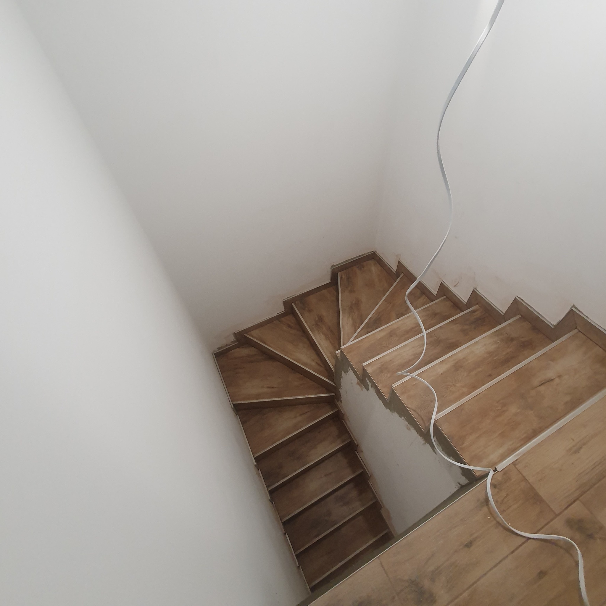 Wewnętrzne schody wyłożone płytkami imitującymi drewno, widok z góry podczas montażu, widoczne białe listwy wykończeniowe i kabel elektryczny zwisający ze ściany.