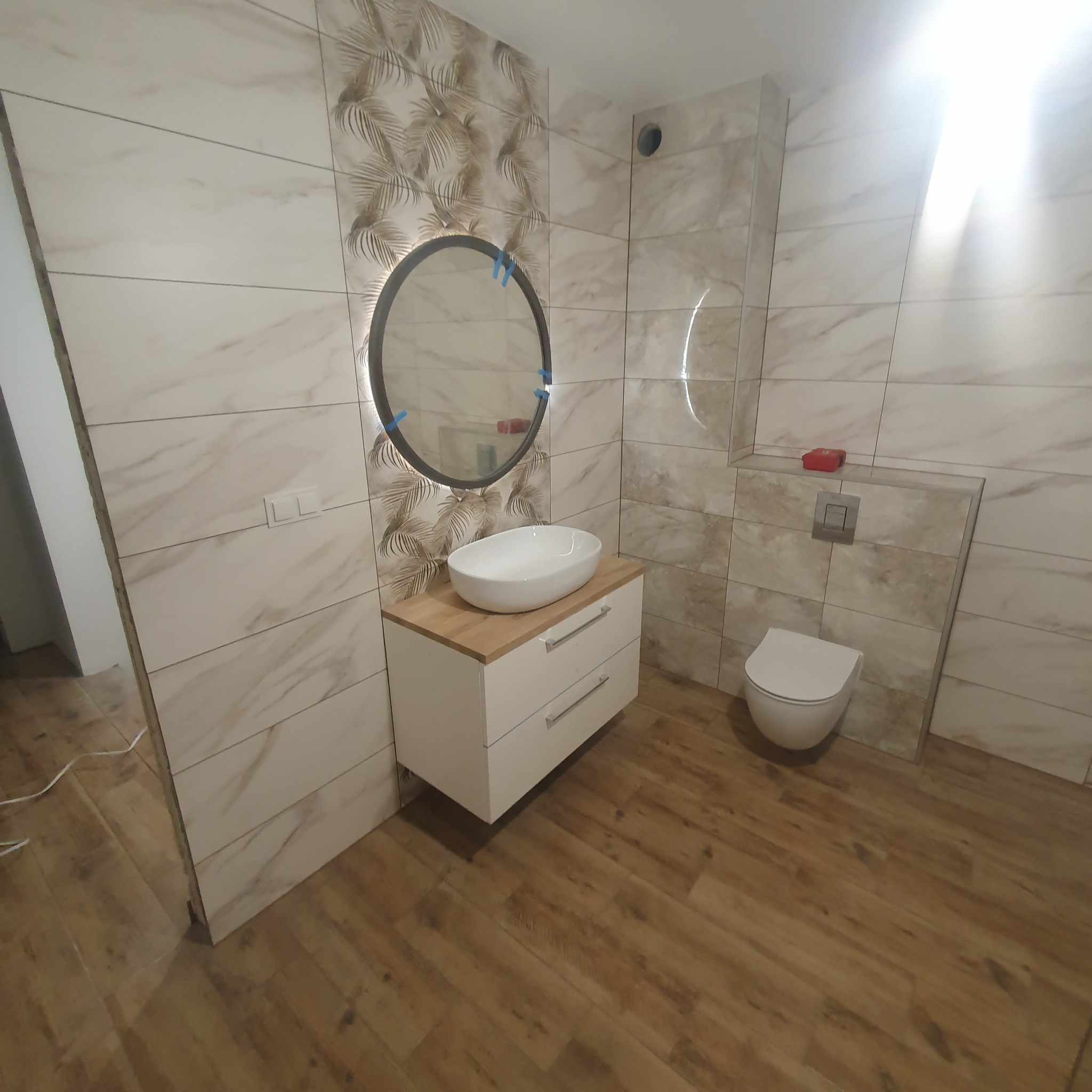 Wykończona łazienka z beżowymi płytkami imitującymi marmur, okrągłym lustrem z podświetleniem LED, białą szafką pod umywalką z drewnianym blatem i podwieszaną toaletą.