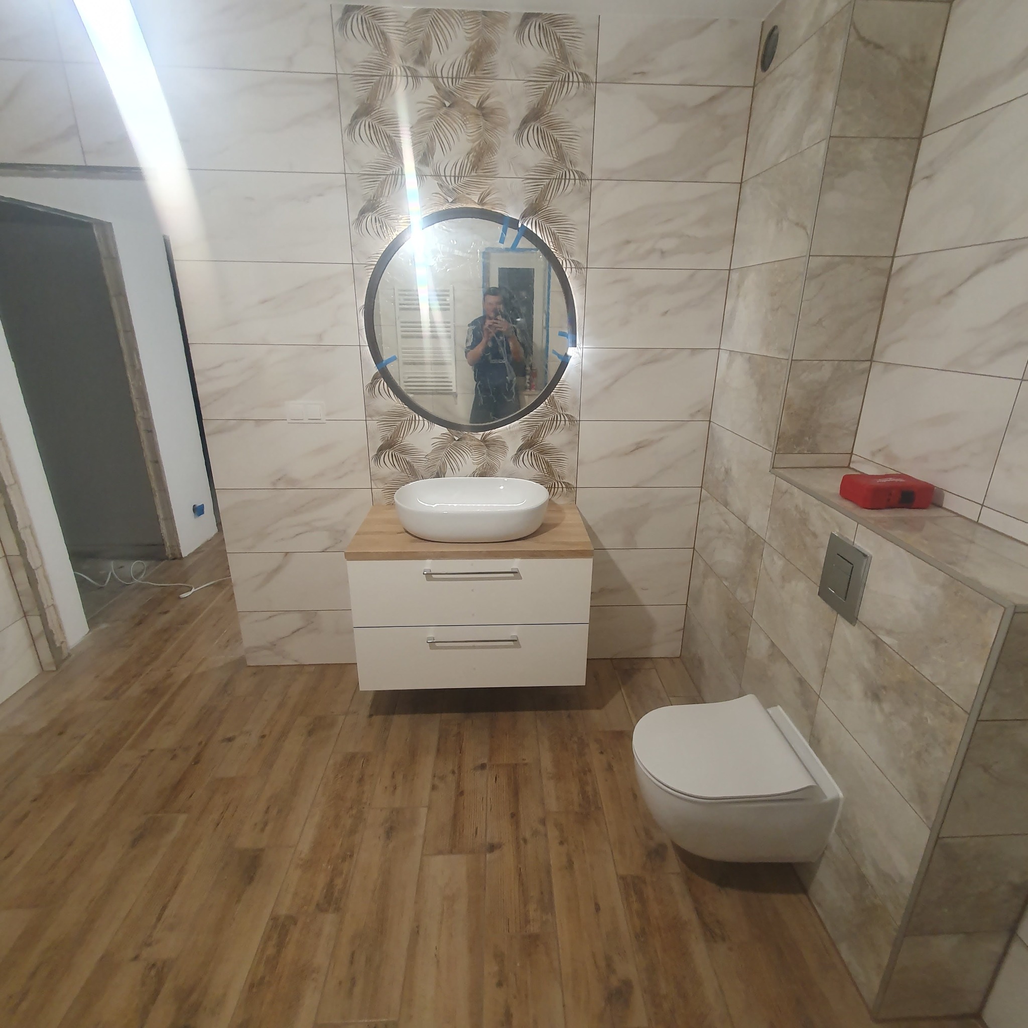 Wykończona łazienka z beżowymi płytkami imitującymi marmur, drewnopodobną podłogą, owalnym lustrem z podświetleniem, białą umywalką nablatową na szafce z drewnianym blatem i podwieszaną toaletą...