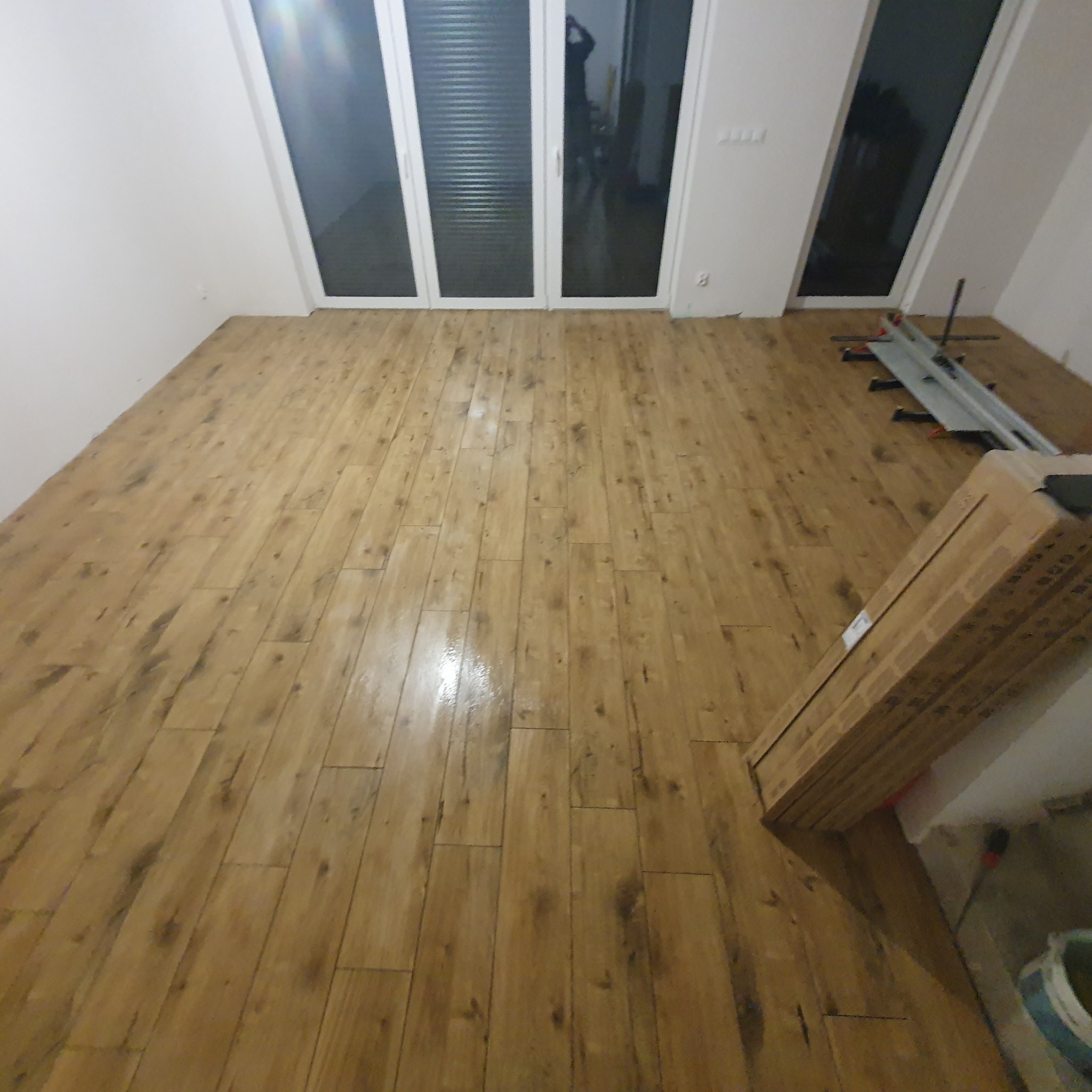 Ułożona podłoga z paneli drewnopodobnych w jasnym odcieniu, widoczne narzędzia do cięcia paneli i paczki paneli czekające na montaż, duże okno balkonowe w tle.