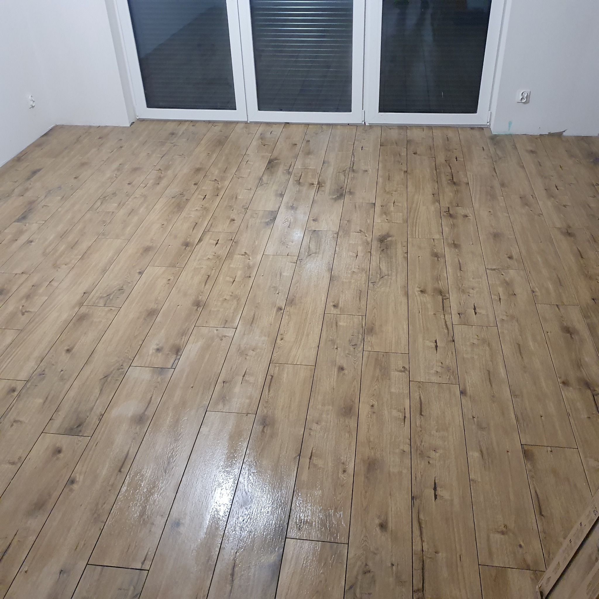 Nowo ułożona, błyszcząca podłoga z paneli imitujących drewno, widoczna przy białych drzwiach balkonowych z żaluzjami.