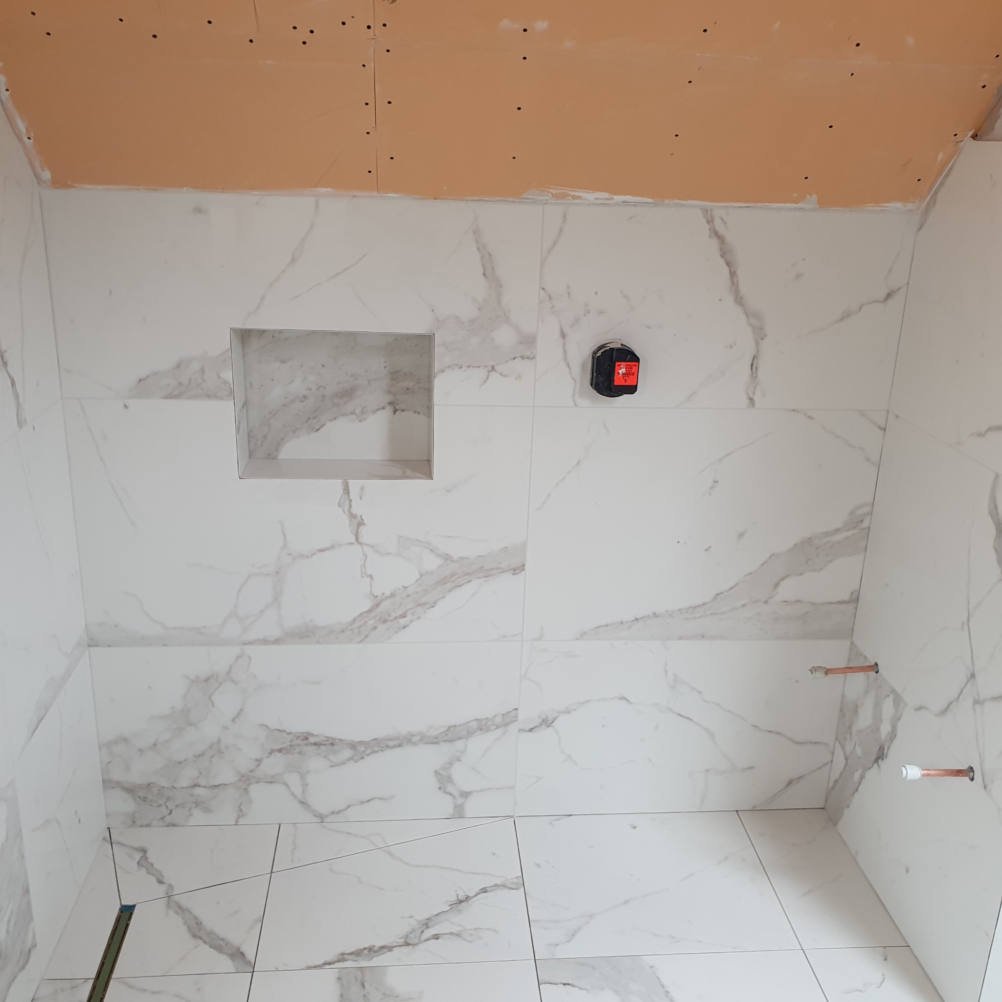 Wnętrze łazienki w trakcie remontu, ściany i podłoga wyłożone białymi płytkami imitującymi marmur, widoczna wnęka w ścianie i niezabudowana płyta karton gips na suficie, poziomica laserowa na ścianie.