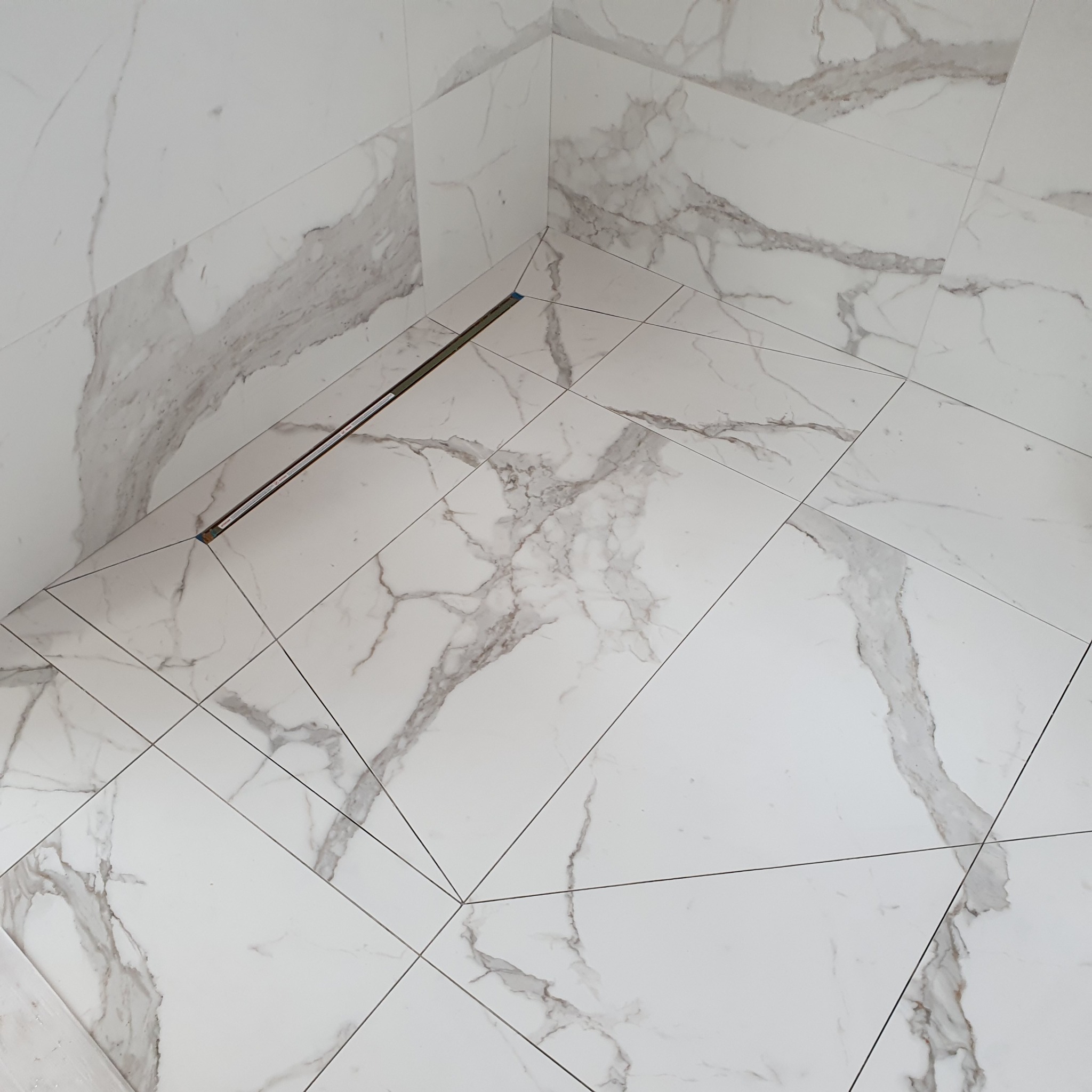 Wykończona łazienka z białymi, marmurkowymi płytkami na ścianach i podłodze, z odpływem liniowym w rogu kabiny prysznicowej.