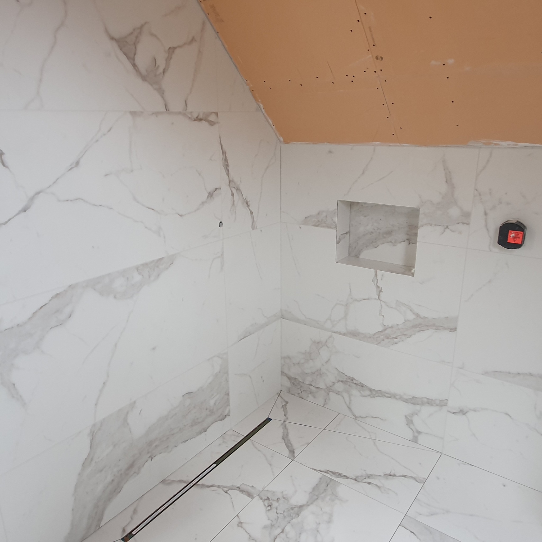 Łazienka w trakcie wykańczania: białe płytki imitujące marmur na ścianach i podłodze, widoczny odpływ liniowy, wnęka w ścianie i fragment sufitu z płyt kartonowo-gipsowych.