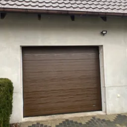 Brązowa segmentowa brama garażowa z poziomymi przetłoczeniami zamontowana w jasnym garażu z dwoma oknami dachowymi i oświetleniem zewnętrznym.