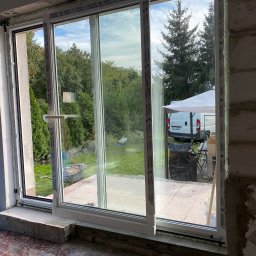 Technika Osłonowa Zubowicz Maksymilian - Montaż okna przesuwnego Salamander z widokiem na ogród, w trakcie prac wykończeniowych. Widoczny wózek budowlany i samochód dostawczy w tle.