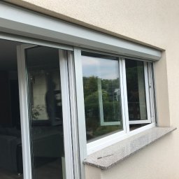 Technika Osłonowa Zubowicz Maksymilian - Białe okno PCV z roletą zewnętrzną, częściowo otwarte, widok z zewnątrz. Na parapecie leży wiertło. Widoczne odbicie zieleni w szybach.