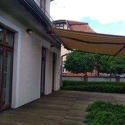 Technika Osłonowa Zubowicz Maksymilian - Beżowo-brązowa markiza tarasowa zamontowana przy budynku z drewnianym tarasem. Widoczne okna z brązowymi ramami i zadbana zieleń w tle.