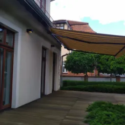Beżowo-brązowa markiza tarasowa zamontowana przy budynku z drewnianym tarasem. Widoczne okna z brązowymi ramami i zadbana zieleń w tle.