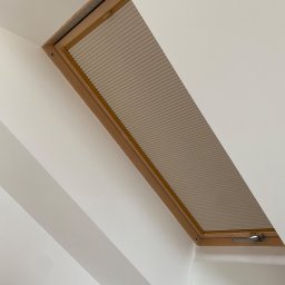 Technika Osłonowa Zubowicz Maksymilian - Okno dachowe z drewnianą ramą i zamontowaną roletą plisowaną w jasnym odcieniu, widoczne z perspektywy wnętrza, przy białej ścianie.