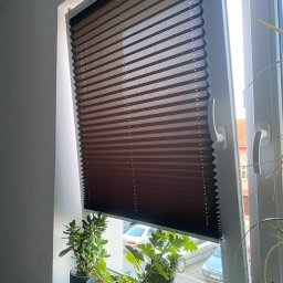 Technika Osłonowa Zubowicz Maksymilian - Ciemnobrązowa plisa okienna zamontowana w białym oknie, częściowo zasłonięta. Na parapecie doniczkowe rośliny. Widok na ulicę przez otwarte okno.
