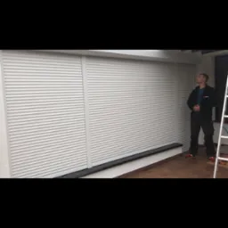 Pracownik w ciemnym ubraniu i czerwonych butach stoi przed trzema białymi roletami zewnętrznymi, spoglądając w górę. Obok stoi biała drabina.