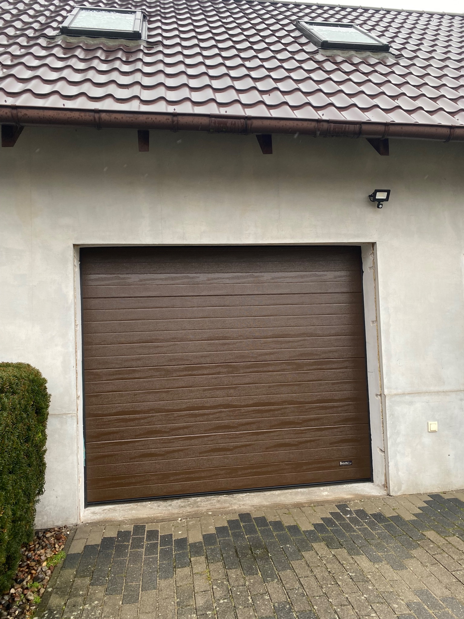 Brązowa segmentowa brama garażowa z poziomymi przetłoczeniami zamontowana w jasnym garażu z dwoma oknami dachowymi i oświetleniem zewnętrznym.