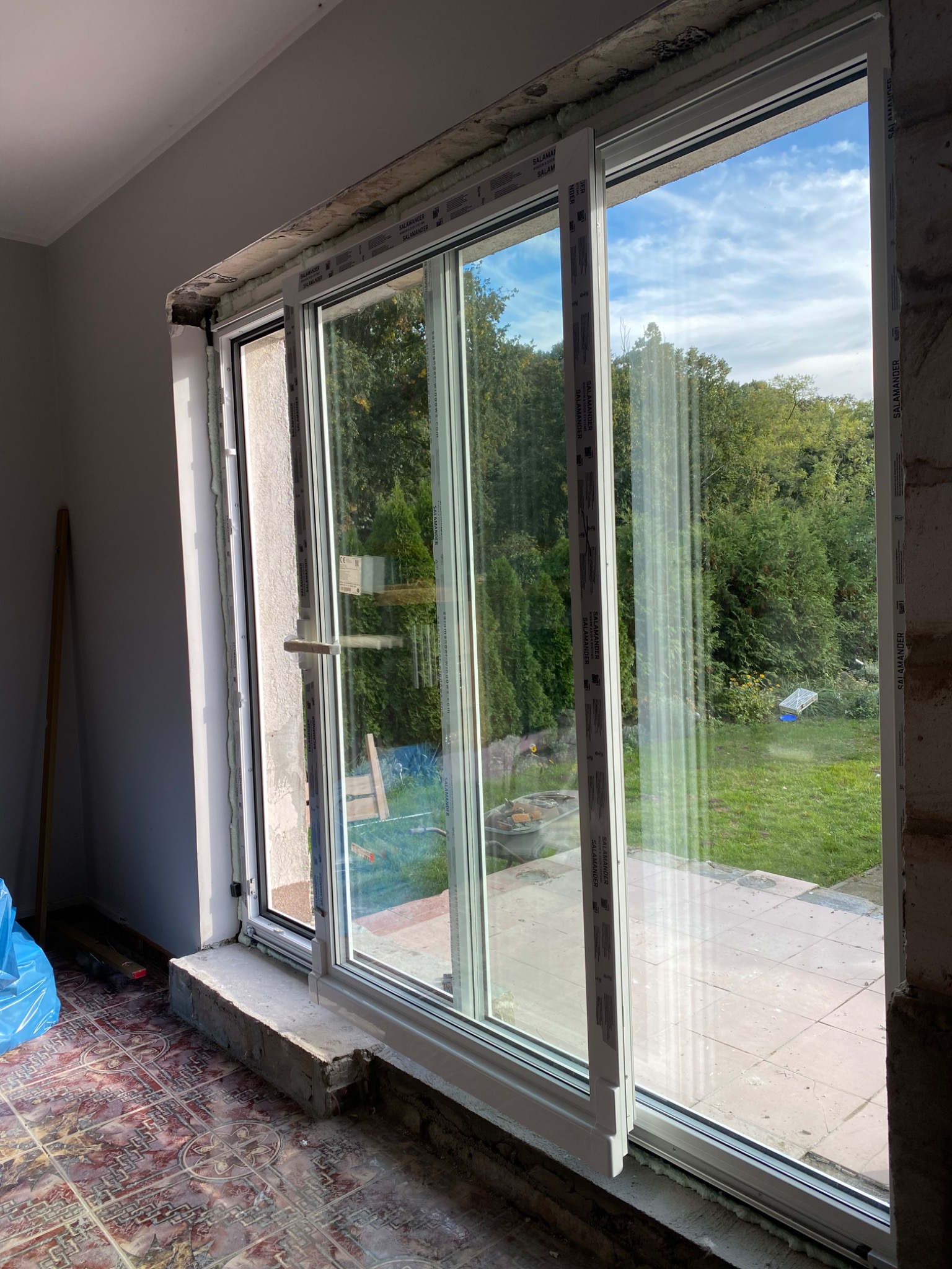 Montaż okna przesuwnego z profili Salamander. Widok na ogród przez duże przeszklenie. Nierówne wykończenie muru i podłogi wskazuje na etap prac wykończeniowych.