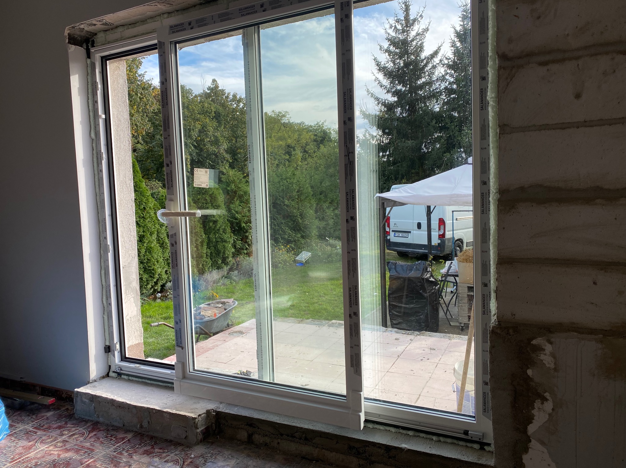 Montaż okna przesuwnego Salamander z widokiem na ogród, w trakcie prac wykończeniowych. Widoczny wózek budowlany i samochód dostawczy w tle.