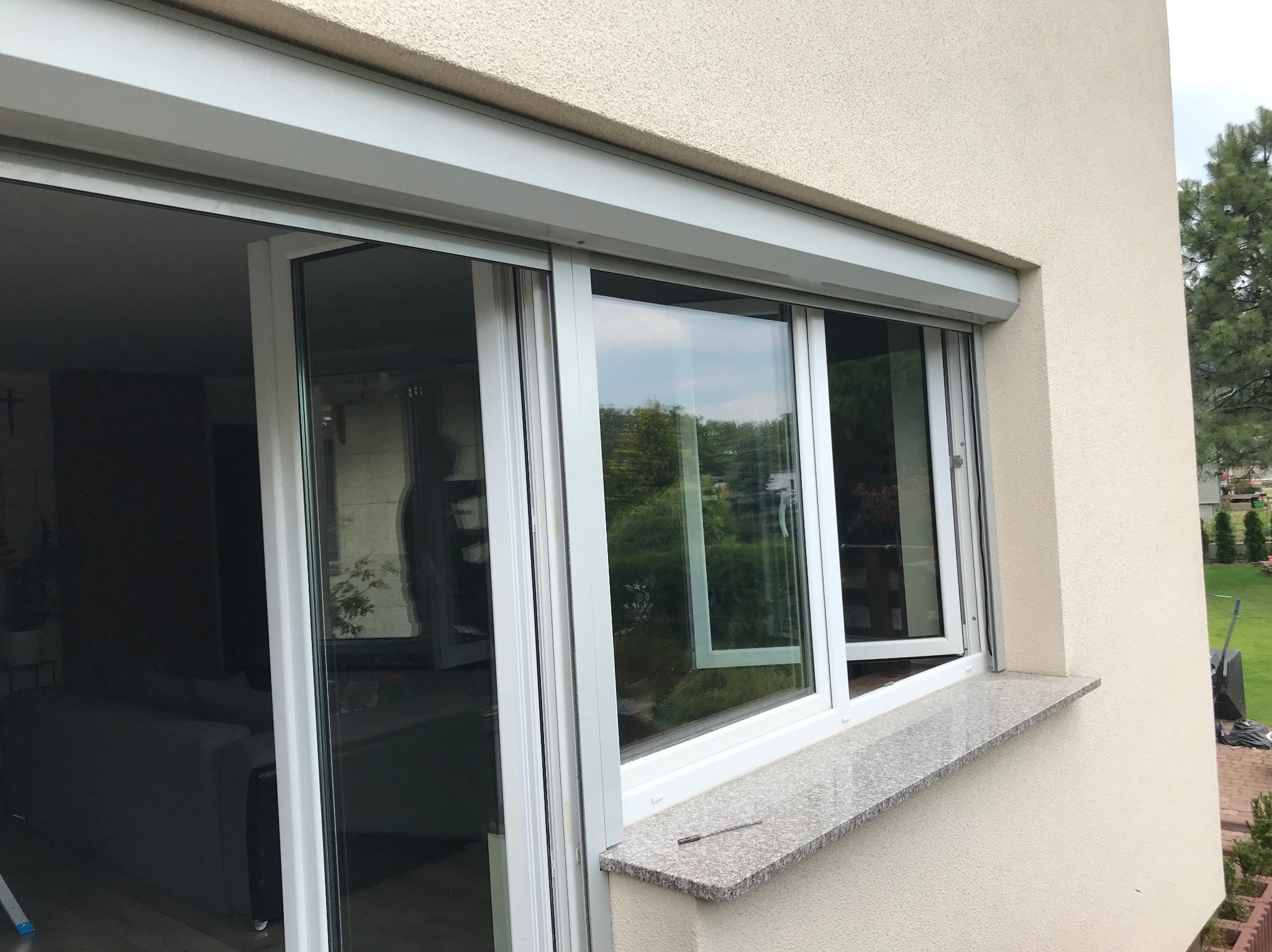 Białe okno PCV z roletą zewnętrzną, częściowo otwarte, widok z zewnątrz. Na parapecie leży wiertło. Widoczne odbicie zieleni w szybach.