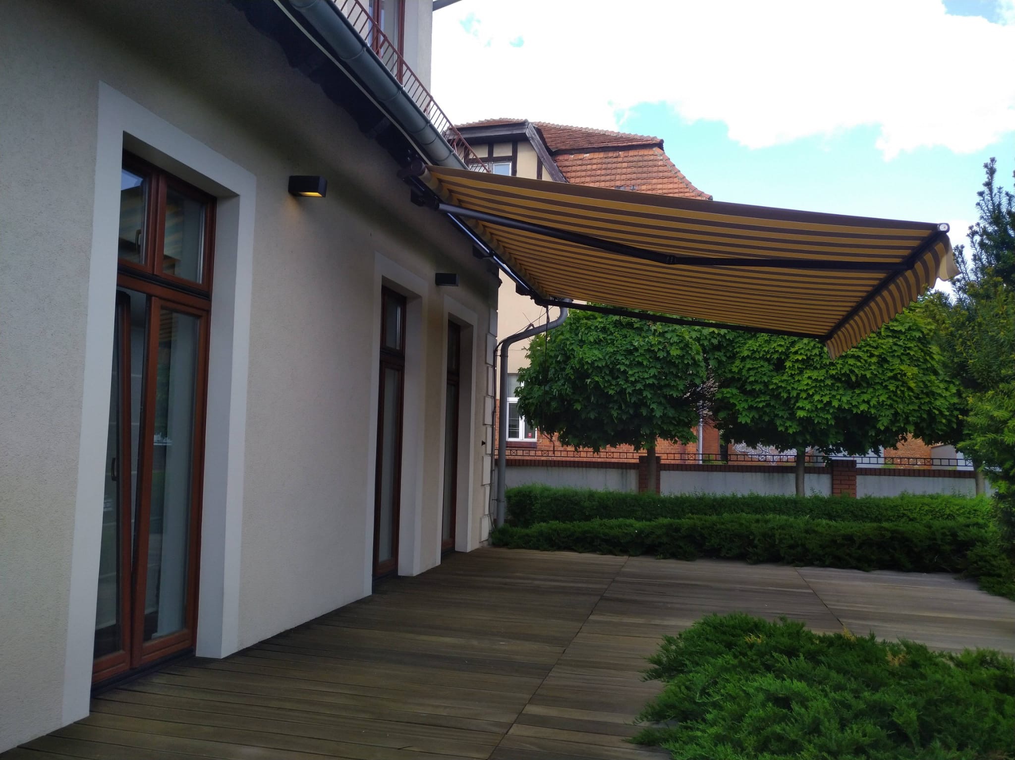 Beżowo-brązowa markiza tarasowa zamontowana przy budynku z drewnianym tarasem. Widoczne okna z brązowymi ramami i zadbana zieleń w tle.