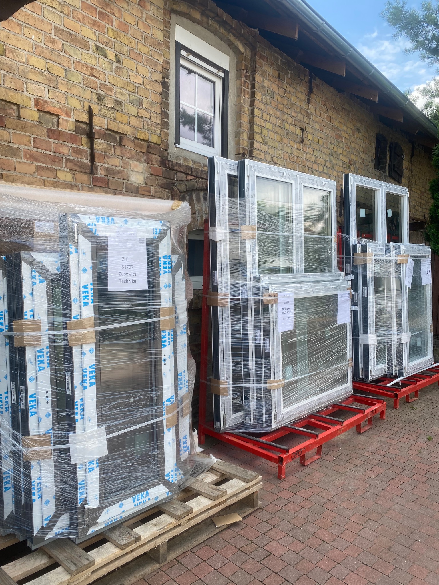 Nowe okna PCV marki Veka, zapakowane w folię, przygotowane do montażu na tle ceglanego budynku. Widoczne etykiety z danymi technicznymi i numerem zlecenia.