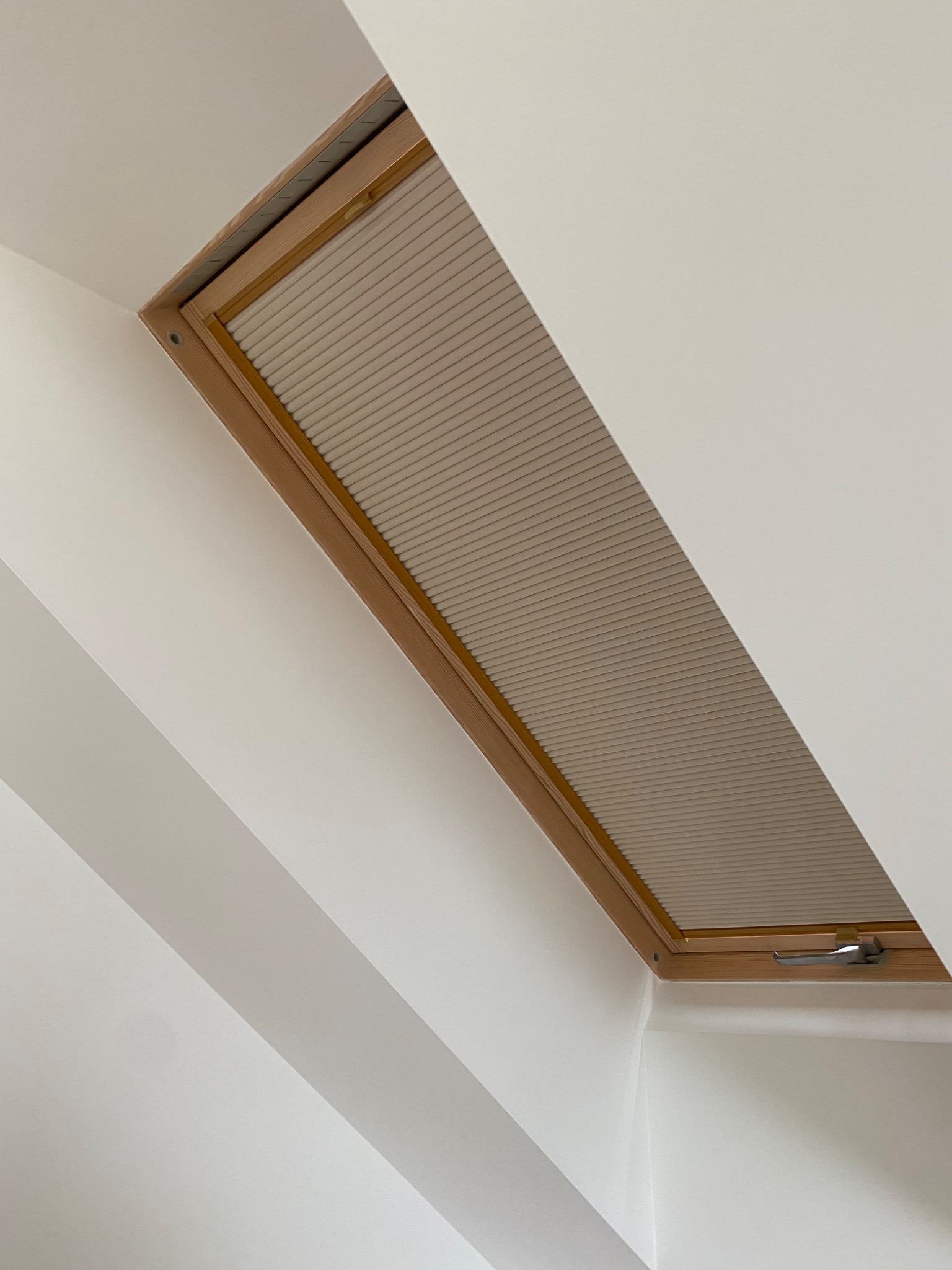 Okno dachowe z drewnianą ramą i zamontowaną roletą plisowaną w jasnym odcieniu, widoczne z perspektywy wnętrza, przy białej ścianie.