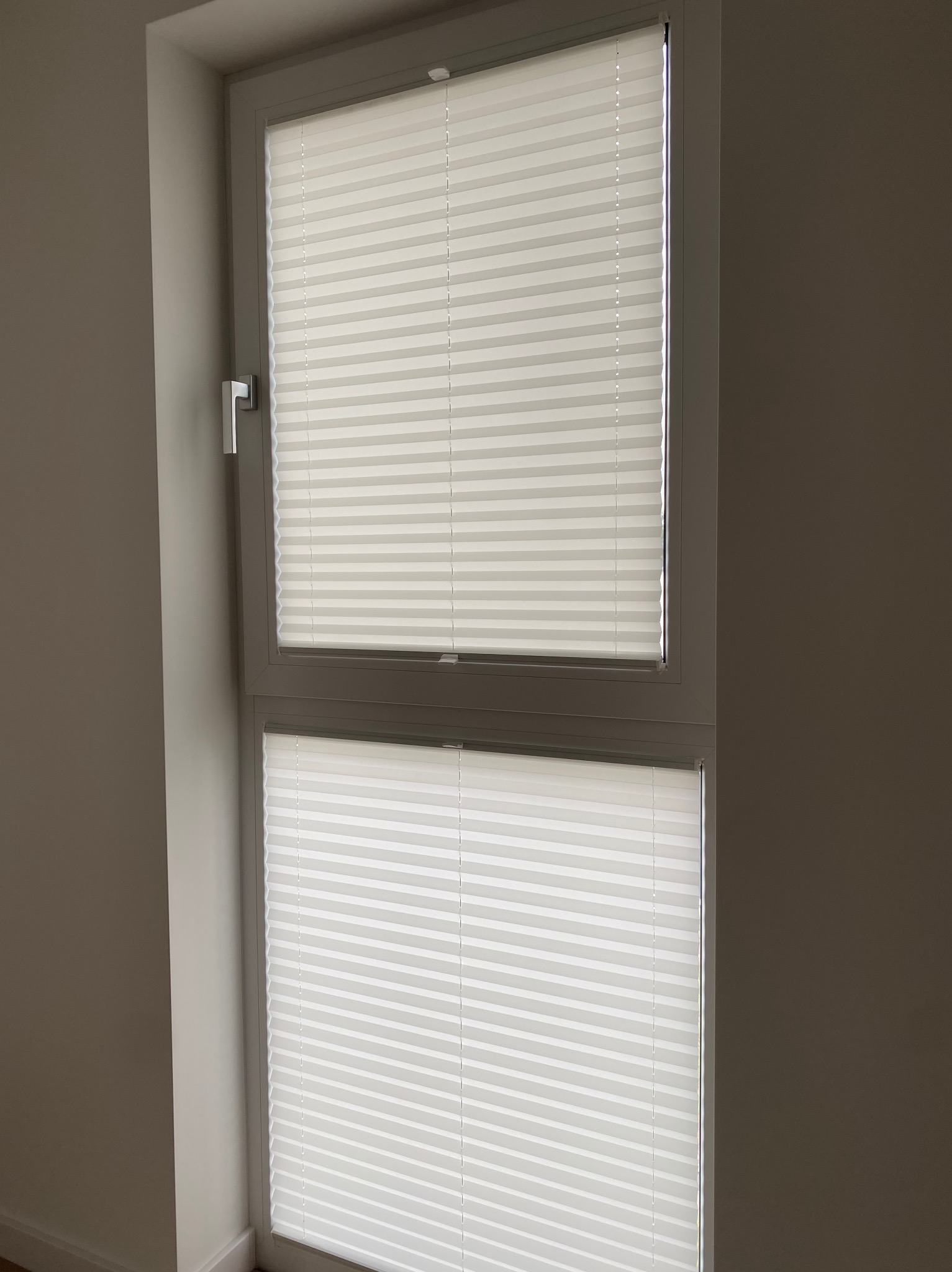 Białe, plisowane rolety okienne zamontowane w dwuczęściowym oknie PCV, widoczne z bliska. Minimalistyczny design, jasne wnętrze.