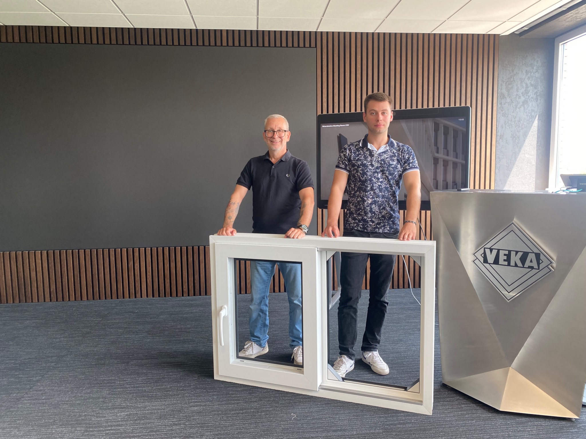 Dwóch mężczyzn prezentuje białe, przesuwne okno PCV marki Veka w nowoczesnym wnętrzu biurowym. Widoczny srebrny pulpit z logo firmy Veka.