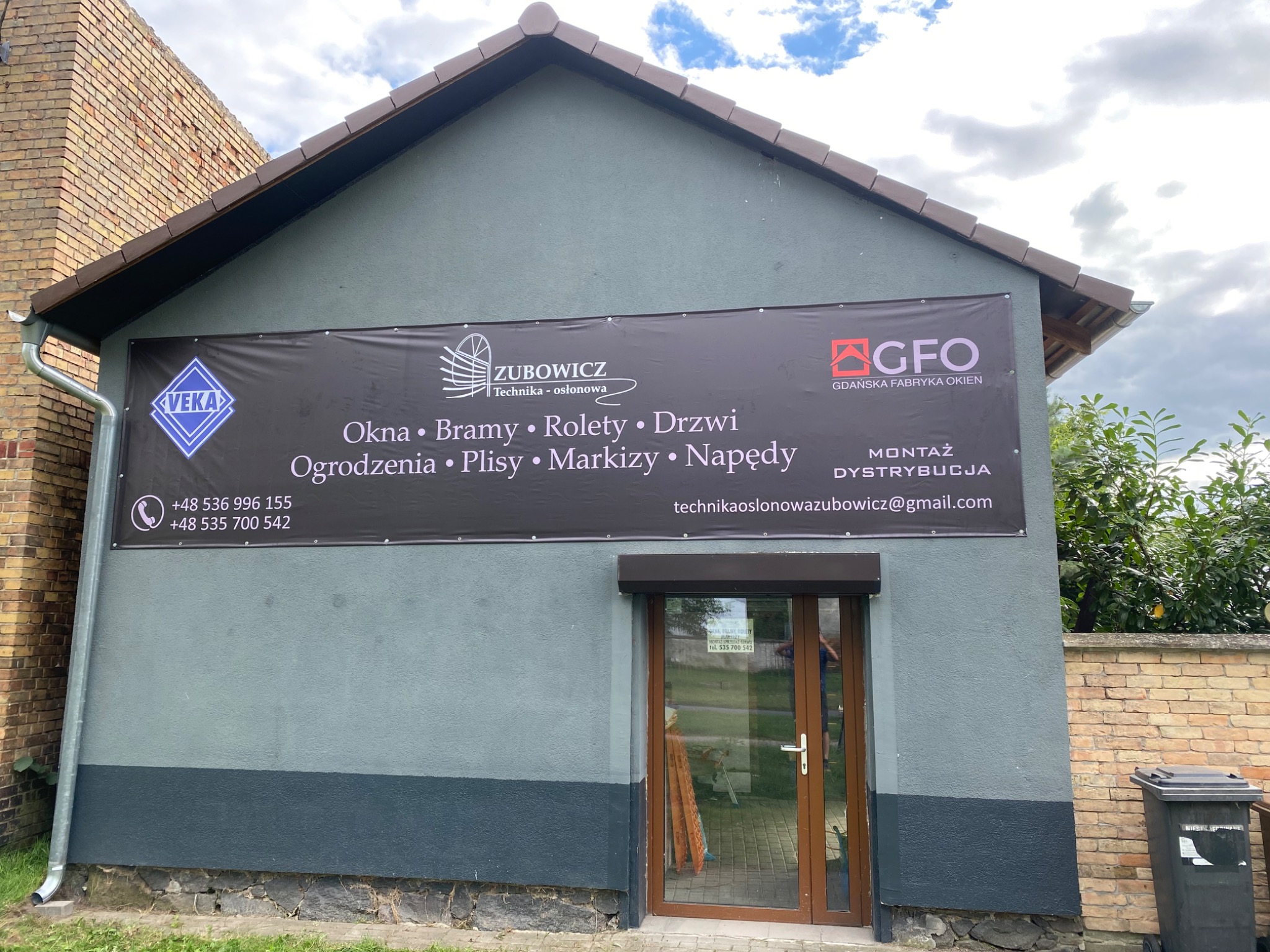 Fasada budynku z szyldem firmy 'Zubowicz' oferującej okna, bramy, rolety i inne. Widoczna brama wejściowa z roletą zewnętrzną. Szyld z logo Veka i GFO.
