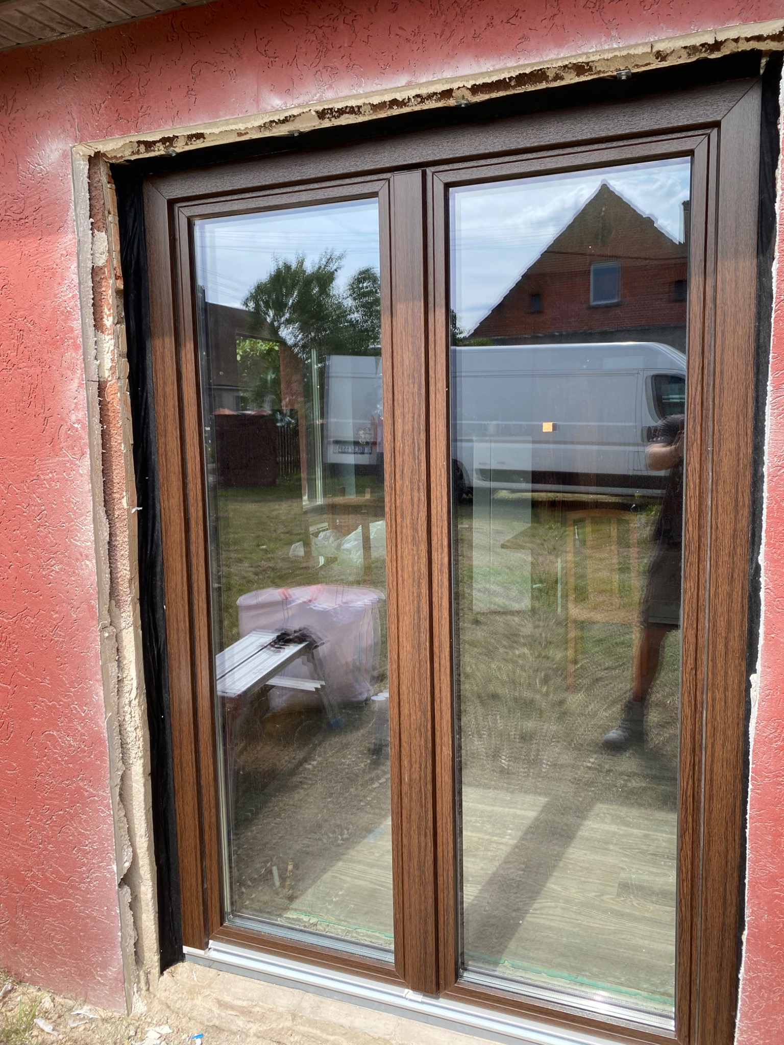 Montaż okna dwuskrzydłowego z brązową ramą w surowym otworze ściennym. Widoczna warstwa izolacji i elementy montażowe. W tle odbicie budynku i samochodu dostawczego.