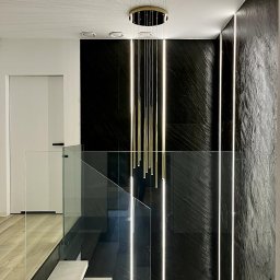 MSN Dawid Lorenc - Nowoczesne wnętrze z podświetlaną ścianą z czarnego kamienia, szklaną balustradą schodów i designerską lampą wiszącą z cienkimi, złotymi prętami.