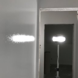 MSN Dawid Lorenc - Świeżo pomalowana, gładka ściana oświetlona lampą roboczą na statywie, widoczny niedokończony otwór drzwiowy z surowym wykończeniem.