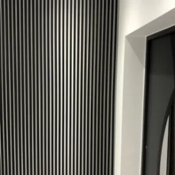 Nowoczesna ściana z pionowych, czarno-białych listew dekoracyjnych, przylegająca do białej ściany z widocznym fragmentem czarnej ościeżnicy drzwiowej.